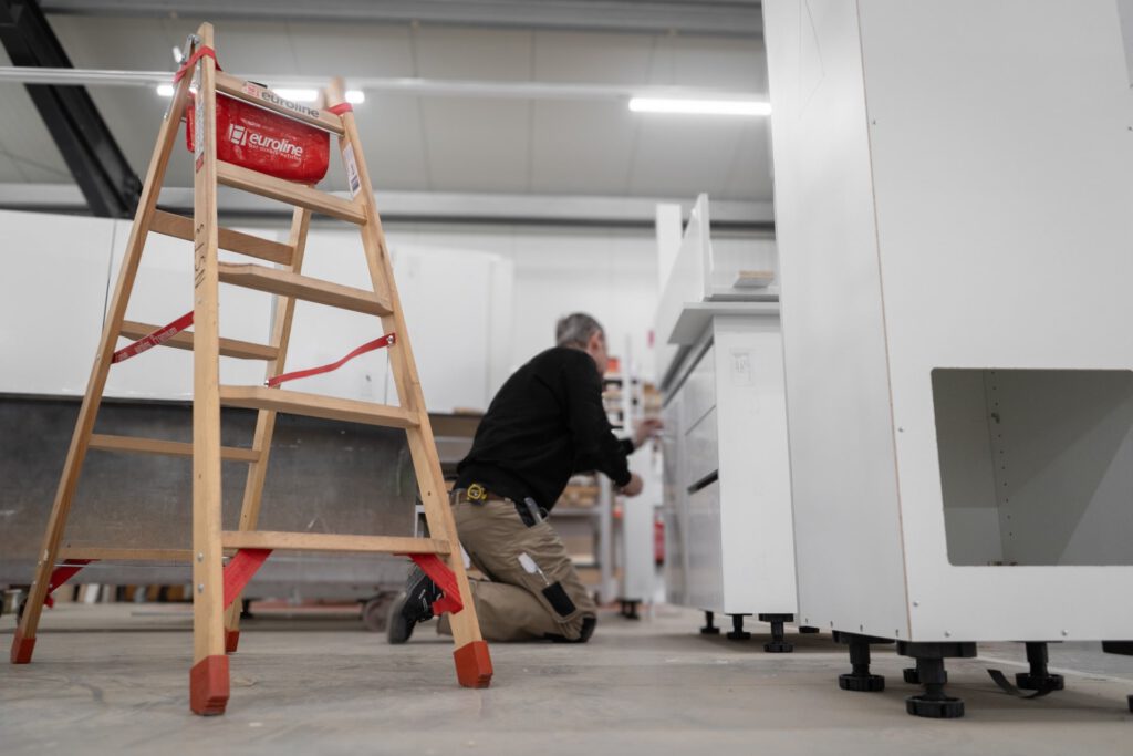 Ein Mitarbeiter arbeitet an einem weißen Möbelstück, während eine Holzleiter im Vordergrund steht.